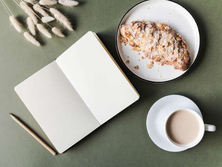 Open notebook with blank sheets, croissant and cup with coffee on green background. Top view.