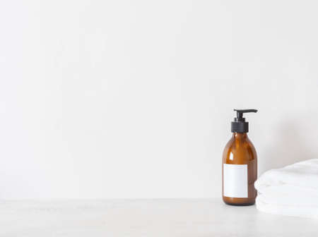 Cosmetic amber glass bottle with white lable and white towels on white background. Front view. Minimal bath background. Copy spaceの写真素材
