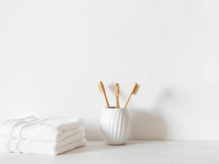 Minimal bath background with bamboo toothbrushs in ceramic glass and white towels on white background. Front view. Copy spaceの写真素材