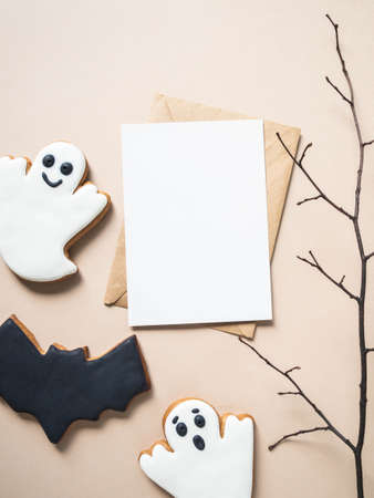 Empty white postcard and craft envelope and Halloween gingerbread cookies - bat and ghosts on beige background. Halloween concept. Top view. Copy space