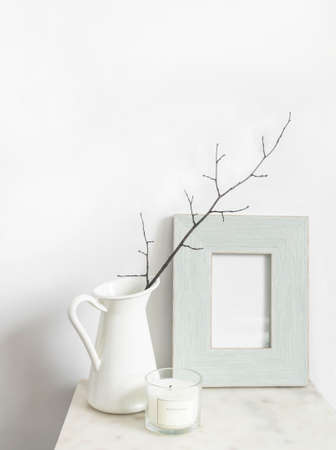 White still life. Scented candle and empty photo frame mockup on marble table. Vase with a branch. Scandinavian interior design. Front view. Copy spaceの写真素材