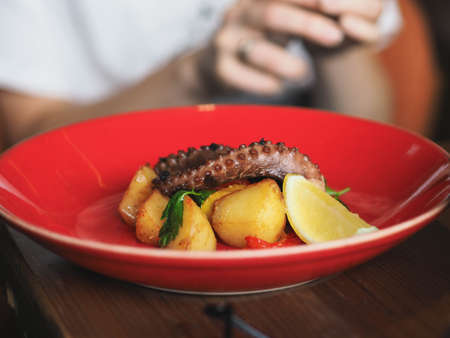 Fried octopus with potatoes on red plate in restaurant. Hands of young uncertain woman with cutlery. Front viewの写真素材