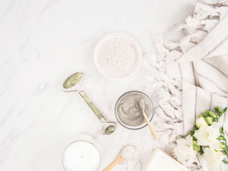 Homemade skincare concept. Organic blue clay for beauty facial mask, crystal massager, candle, cotton towel and white flowers on marble background. top view. copy spaceの写真素材