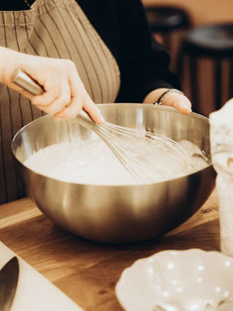 Indeterminate woman mixed batter with whisk in large metal bowl. Home baker whisking batter preparing delicious mealの写真素材