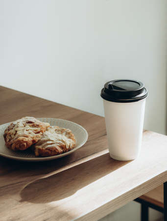 Cup of coffee to go in white paper cup and croissants in sunlight on wood table. Morning coffee in cafe Place for text.の写真素材