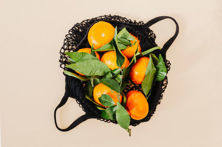 Black grocery eco-mesh cotton bag with fresh orange tangerines with leaves on a beige pastel background. top view. flat lay.の写真素材