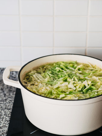White ceramic frying pan with chopped cabbage in broth on the stove. Stage of preparation of vegetable soupの写真素材