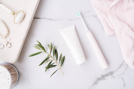 Top view of white plastic tube without label, electric toothbrush and olive branch on white bath...