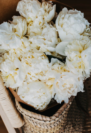 Bouquet of white peony flowers in a wicker bag on brown chair in room. Top viewの写真素材