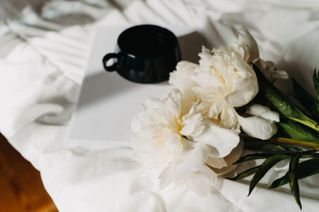 Bouquet of white peony flowers, magazine, cup of tea on white bad in sun light. Top view. Copy spaceの写真素材