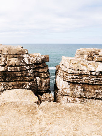 A dramatic coastal scene showing a rugged cliff split into two sections, revealing the ocean in the background. The textured rock formations frame the tranquil sea under a soft, cloudy skyの写真素材