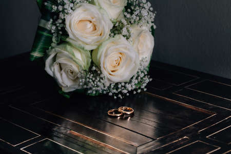 Two golden wedding rings on retro wooden table with white rose bouquet flowers in background