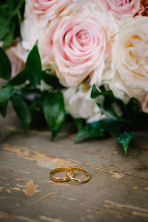 Close up view two golden wedding rings on retro wooden table with white rose bouquet flowers in backgroundの写真素材