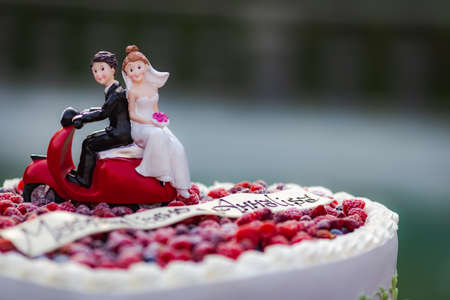 Close up decorated fresh wedding cake with two figures of bride and groom on red scooterの写真素材