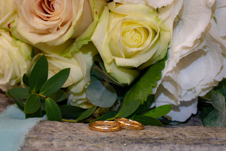 Two golden rings with engraved bride and groom names lay on surface by rose flower bouquetの写真素材
