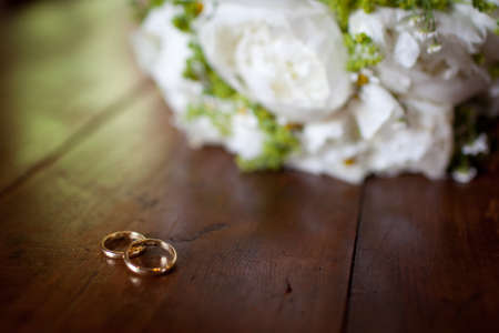 To golden wedding rings lie on wood flooring by beautiful white rose bouquet out of focus. Weds ring blank space retro backgroundの写真素材