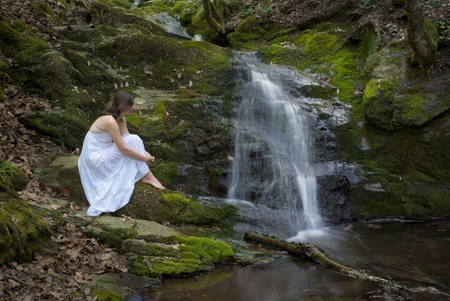 Beautiful young woman in a white dress contemplates a waterfall in the middle of a forestの写真素材