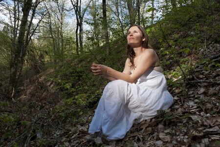 Young woman wearing a white dress sitting on the ground in a forestの写真素材