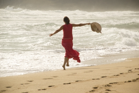 Beautiful young happy woman wearing red dress playing with the waves on a sandy beachの写真素材