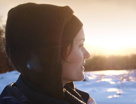 Young beautiful woman with a hat and a hood walking in the snow at sunsetの写真素材