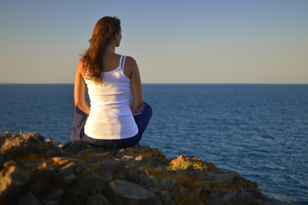 Beautiful young woman sitting on a rock contemplating the blue evening sea.の写真素材