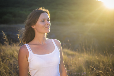 Beautiful young woman looking at the view in a natural field with the golden light of the setting sun shinning onto her.の写真素材