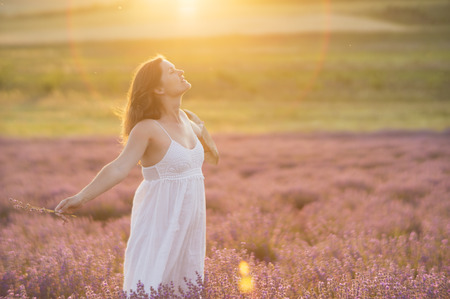 Beautiful young woman with a white dress and a straw hat standing in the middle of a lavender field at in the golden light of the sunset praising the beauty of life.の写真素材