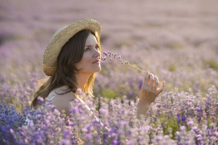 Beautiful young woman wearing a straw hat, holding a bunch of lavender flowers, enjoying their fragrance sitting in the middle of a lavender field at sunset.の写真素材