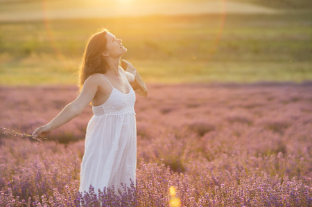 Beautiful young woman with a white dress and a straw hat standing in the middle of a lavender field at in the golden light of the sunset praising the beauty of life.の写真素材