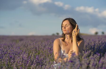 Beautiful young woman holding a bunch of lavender flowers enjoying their fragrance in the middle of a lavender field at sunset.の写真素材