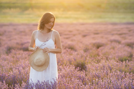 Beautiful young woman wearing a white dress standing in a moment of peace and serenity  in a middle of a lavender field under the golden light of sunset.の写真素材