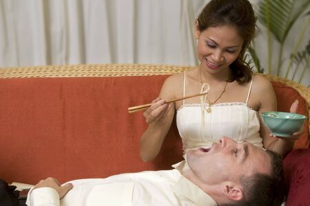 Asian woman feeding caucasian man noodles while  laying on a sofaの写真素材