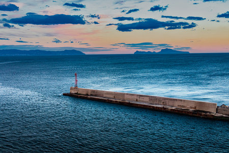 The image shows a long, gray concrete pier extending into the ocean. The pier is flanked by a tall red communication tower. A distant coastline, featuring mountains, is visible across the water. The sky is overcast with gray clouds, suggesting a cloudy day.の写真素材
