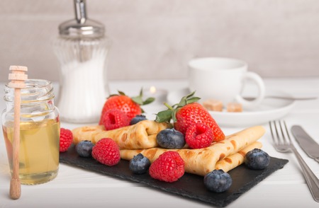 Pancake dish with fresh fruits strawberry blueberry and raspberry on a white wooden table. On the side coffee and honey and sugar.の写真素材