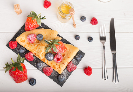 Pancake dish with fresh fruits strawberry blueberry and raspberry on a white wooden table. On the side coffee and honey and sugar.の写真素材