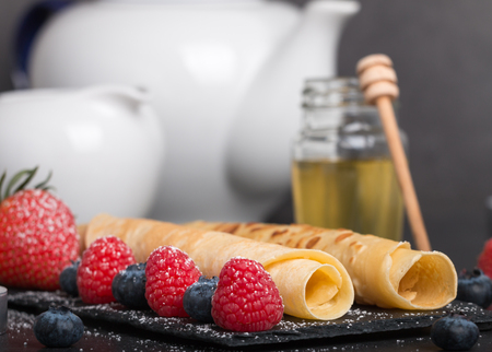 rolled pancakes with fresh strawberryâs and raspberryâs and blueberryâs on a black stone plate on a dark stone table with honey and white sugar and coffee.の写真素材
