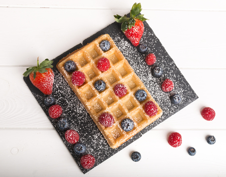 Freshly baked Brussels waffles on a black stone plate with fresh strawberryâs and blueberryâs and raspberryâs on a white wooden table with honey and coffee.の写真素材
