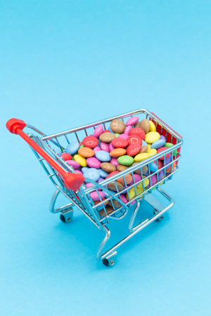 Shopping cart for children's birthday with many colorful smarties sweets in different colors. Blue background, verticalの写真素材