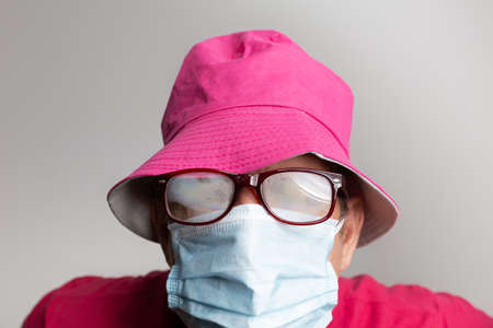 Man with medical mask and fogged glasses and pink hat on grey background