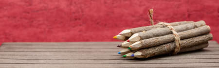 bound crayons made of untreated, real wood with real tree bark on wooden board with red background for students, artists in banner format. Horizontalの写真素材