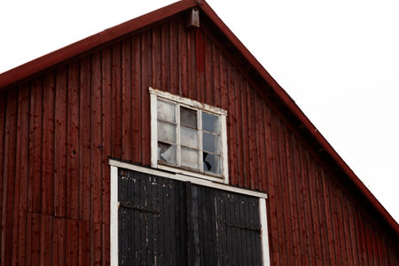 A close-up view of a traditional Scandinavian red wooden barn featuring vertical siding, a black wooden door, and a small white-framed window under the peaked roof against a white sky.の写真素材