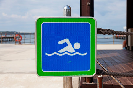 A blue and green swimming sign with a swimmer icon displayed at a wooden pier overlooking a calm body of water with safety equipment visible in the background.の写真素材