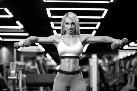 Sexy fitness blonde woman is doing side raise with dumbbells in the gym BWの写真素材