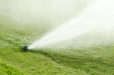 Impact sprinkler on lawn in action. Impulse sprinkler head with out streaming water fountain on artificial green lawn in full sunlight. Long spray radius creates the effect of natural rainfall. Photo.の写真素材