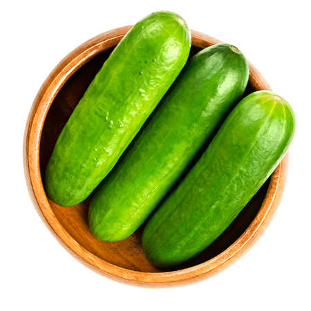 Fresh snack cucumbers in a wooden bowl. Group of small, young and whole cucumber fruits, lined up close together. Cucumis sativus, a vegetable. Close-up, from above, isolated, over white, food photo.の写真素材