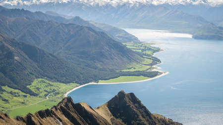 Beautiful vista with dry tussocks,lush green meadows,blue lake and snowy mountains narrow shot made during sunny day on Isthmus Peak summitの写真素材