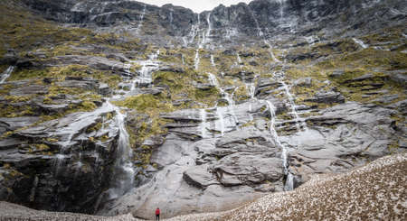 Huge wall of waterfalls with person located on the bottom for scale. Photo taken in Fiordland National Park, New Zealand.の写真素材