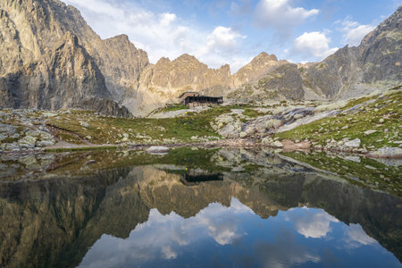 Alpine hut surrounded by mountains reflected in foreground tarn during sunrise, Slovakia, Europeの写真素材