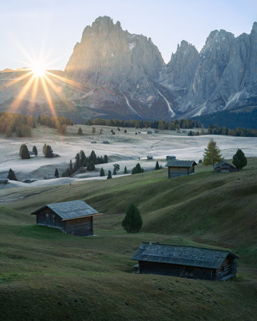 Vertical shot of Sun rising above high alpine pasture with cottages from behind a wall of mountainsの写真素材