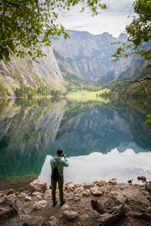 Vertical shot of man taking photo on bank of still beautiful greenish alpine lake, Obersee, Germanyの写真素材